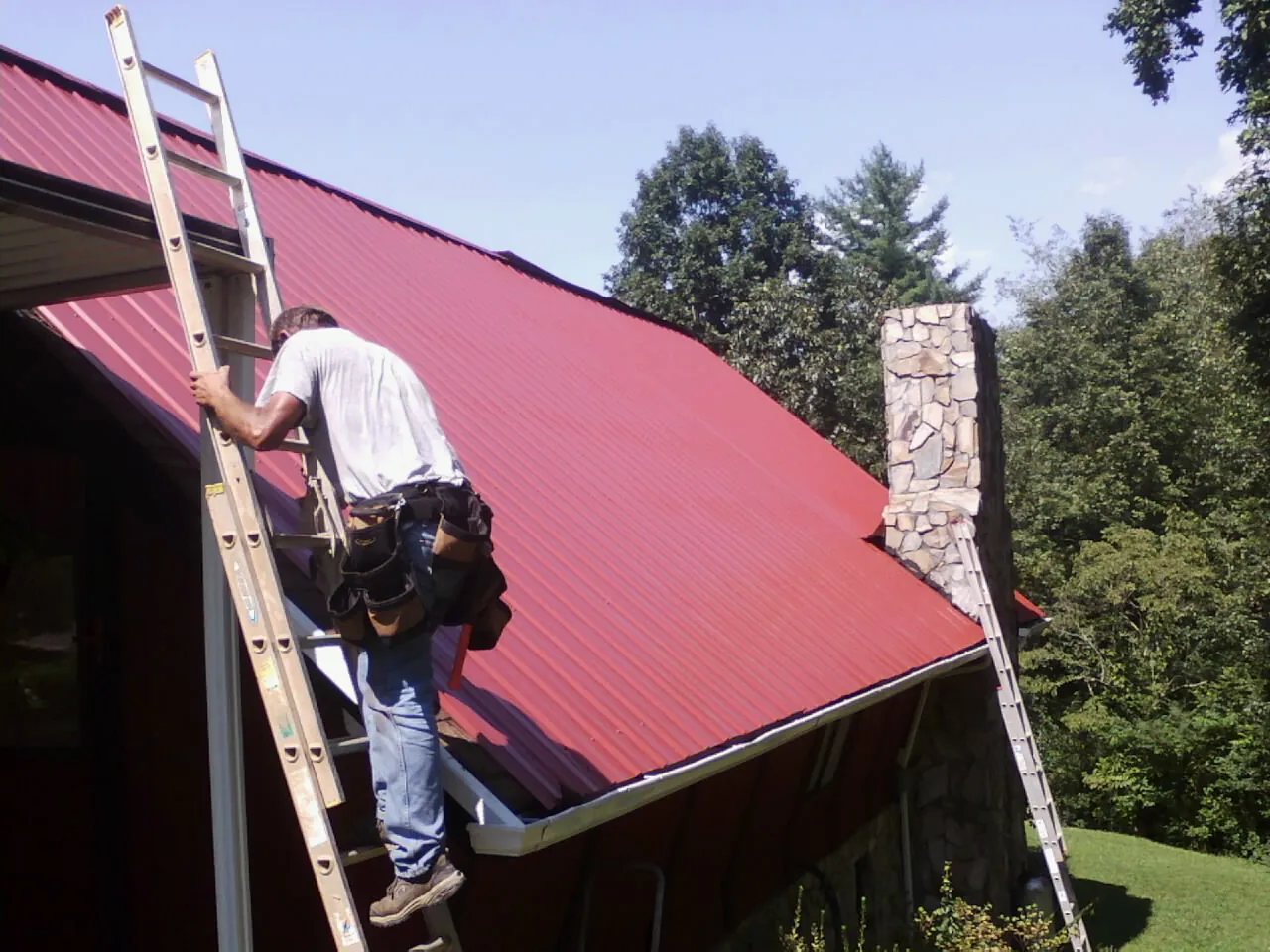 Roofer on a ladder beside a red metal roof and stone chimney — Soffit & Fascia Repair by Roofing Brothers in Mantador